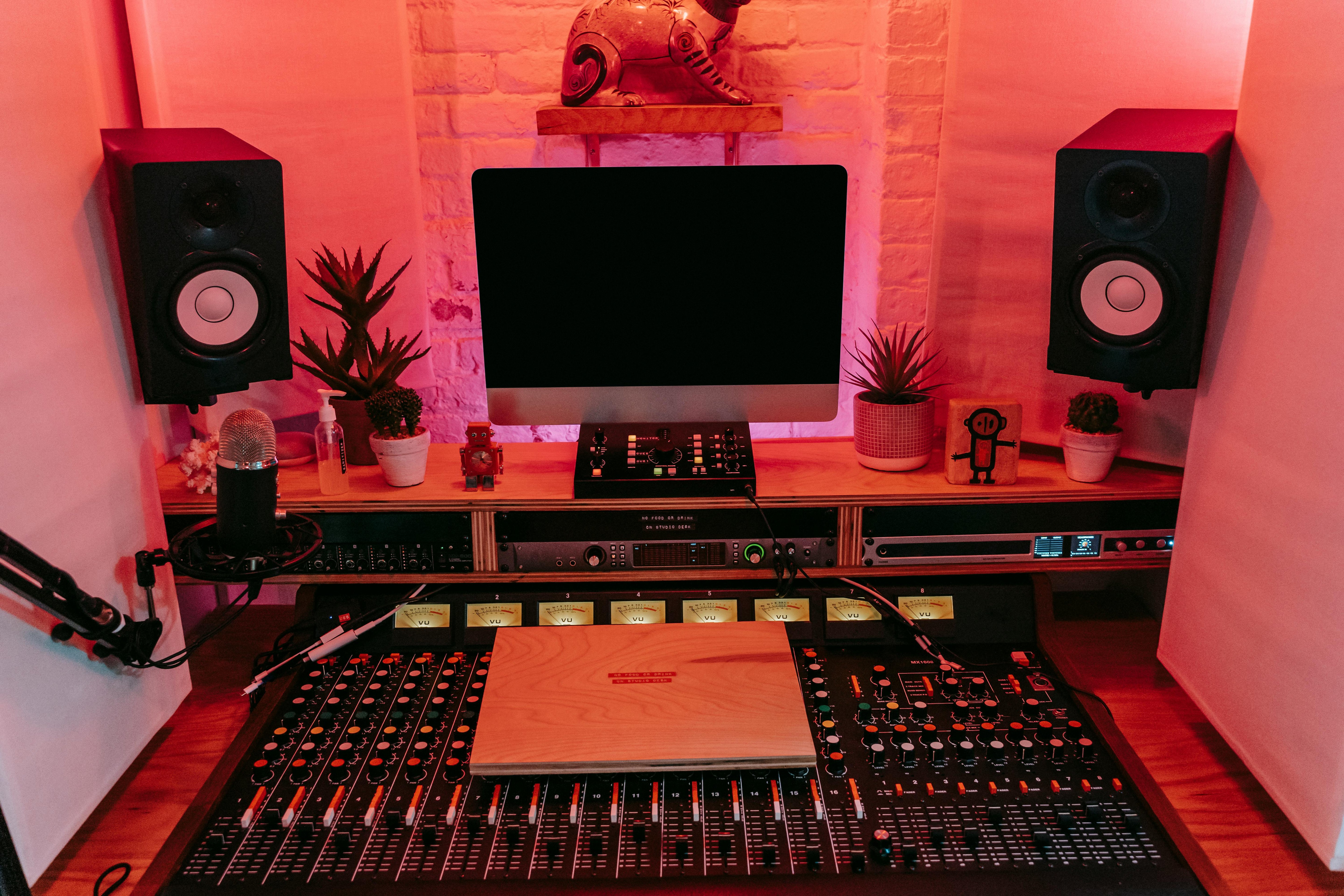 Studio amenajat acasa, cu birou din lemn, microfon pe stativ, monitor audio, ecran de computer si panouri/bureti acustici montati pe perete, intr-un decor modern si cald.
